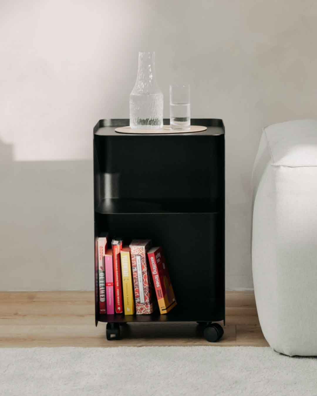 Schwarzer JUNO Rollcontainer in großer Ausführung neben einem Sofa mit Büchern und Glasflasche